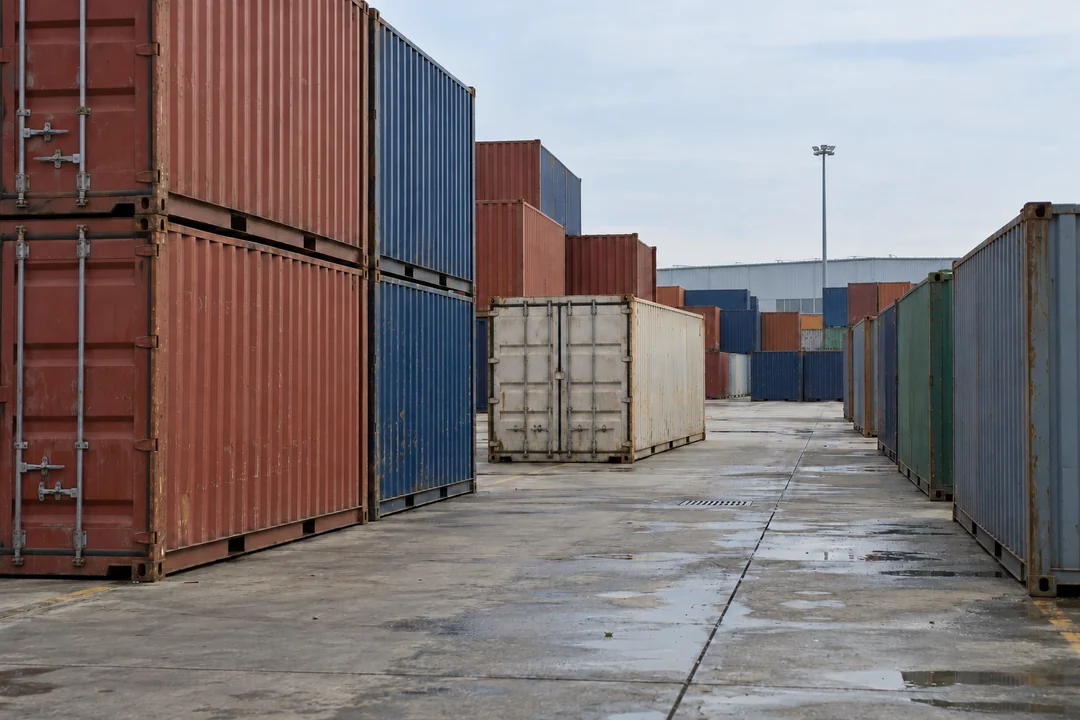 Illustrative image of stacked red and blue shipping containers beside a paved lane and warehouses